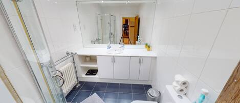 A modern bathroom with a glass shower enclosure, white tiles, a white sink with a mirror above, and a toilet with toilet paper and a bottle of hand soap.
