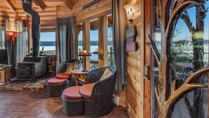 Un acogedor interior de cabaña con chimenea de piedra, muebles de mimbre y amplias ventanas que ofrecen vistas al exterior.