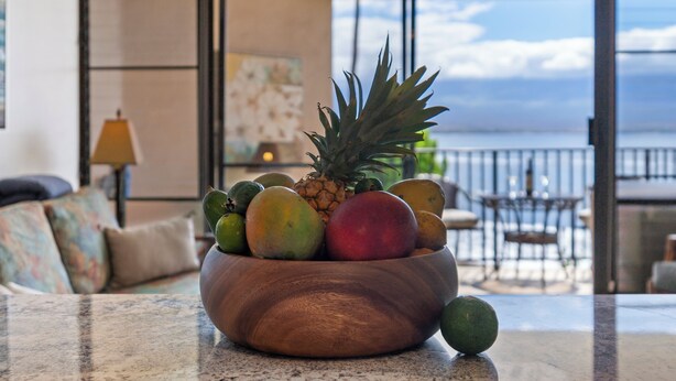 A wooden bowl of fruit on a kitchen counter with a view of a balcony and outdoor seating area. - Relaxing Oceanfront! (Wailuku)