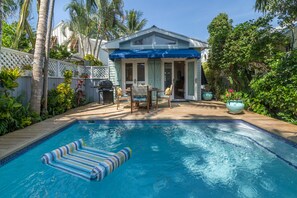 Ein Swimmingpool mit Liegestuhl, eine überdachte Terrasse mit Gartenmöbeln und ein Haus mit blauem Markisenvorhang.