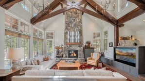 A spacious living room with a stone fireplace, large windows, and a high ceiling with exposed beams. - MONTHLY LEASE - Luxury 4 BR 4 BA Truckee Available March 2022 (Truckee)