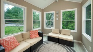 Ein Zimmer mit einer beige farbenen Couch, einer dunklen Wickerbänkchen und einem großen Fenster mit Blick auf Grünflächen.