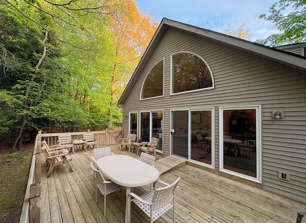 A wooden deck with a table and chairs, large glass doors leading to a covered patio area, and a house with a gabled roof.