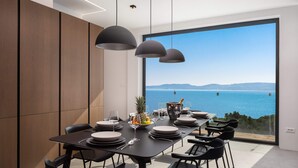 A modern dining area with a large table set for six, a view of the sea, and pendant lighting. - Wind Rose Wellness Villa Maestral (Marušici)