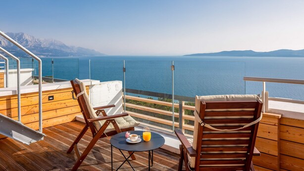 A balcony with wooden flooring, two wooden chairs, a small round table with a cup and orange juice, and a glass railing overlooking a calm sea and distant mountains. - Wind Rose Wellness Villa Maestral (Marušici)