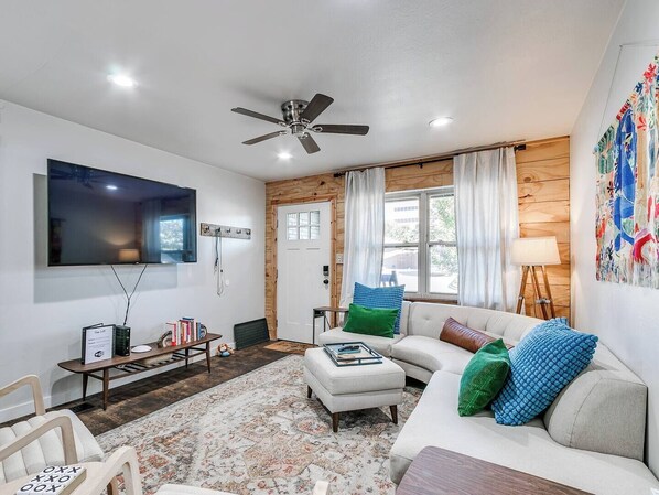 A living room with a flat-screen TV, a ceiling fan, a wooden door, and a window with curtains.