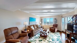 A spacious living room with a large window, a sofa, two armchairs, a coffee table, and a TV.