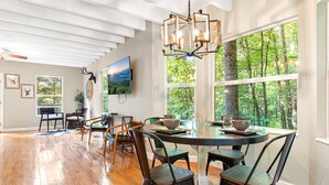 A dining area with a round table, chairs, and a chandelier. There are windows with a view of trees.