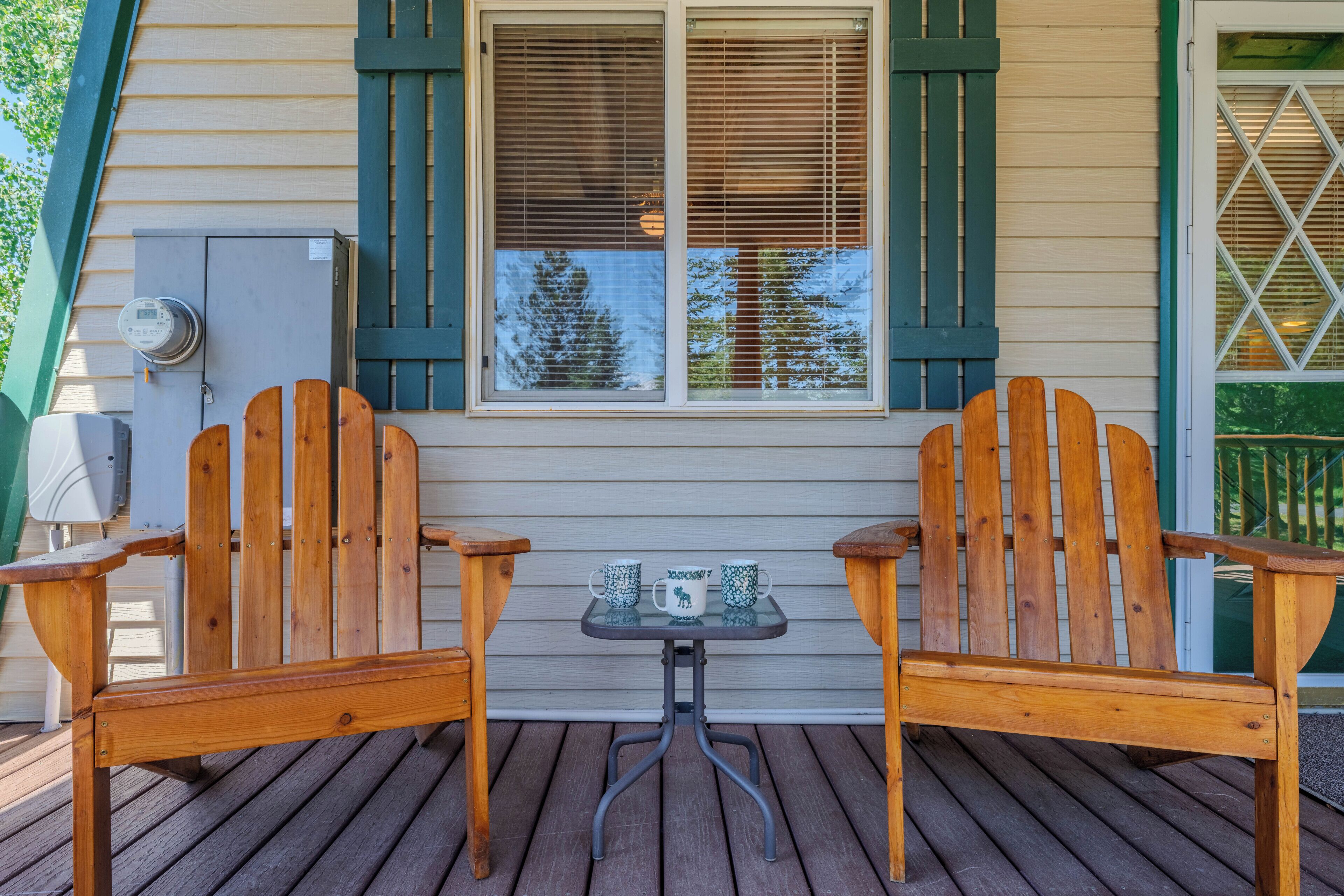 A wooden deck with two Adirondack chairs and a small table, a window with shutters, and an electrical box.