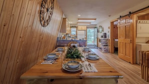 Una mesa de comedor de madera dispuesta con platos, vasos y cubiertos, una rueda decorativa en la pared, y una cocina con armarios y electrodomésticos de madera.