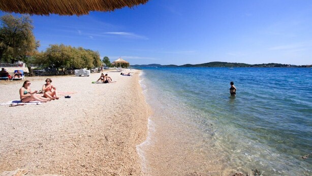 Une plage avec une eau claire et peu profonde, un parasol en chaume et des gens qui se détendent.