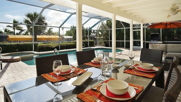 Un espacio de comedor al aire libre con una mesa de vidrio, sillas y vistas a una piscina y palmeras.