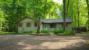 A single-story cabin with a front porch, surrounded by a gravel driveway and lush greenery. - 2 Miles to Good Harbor Beach, Pet Friendly (Cedar)