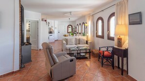 A spacious living area with a sofa, armchair, and a dining table. There are arched windows and a kitchen in the background.