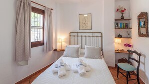 A neatly made bed with a white bedspread, two pillows, and two rolled towels with a logo. There is a window with curtains, a bedside table with a lamp, a chair, a bookshelf with books, and a framed picture on the wall.