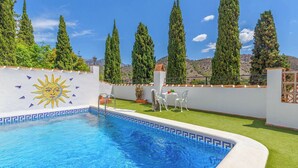 Ein Schwimmbecken mit Sonnendesign, umgeben von einer weißen Mauer mit dekorativer Bordüre. Zwei weiße Stühle und ein kleiner Tisch mit einer Pflanze stehen daneben. Im Hintergrund sind hohe Bäume und ein Berg zu sehen.