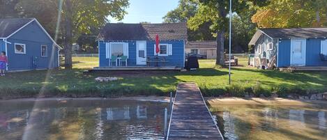 A small blue cabin with a dock extending into a pond, surrounded by grassy areas and trees.