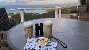 A round table with two glasses of a light-colored beverage, a pair of binoculars, and a plate of food on a balcony overlooking a coastal landscape. - Ultimate USVI Views overlooking Caribbean & Salt River Bay Nat'l EcoPreserve (Christiansted)