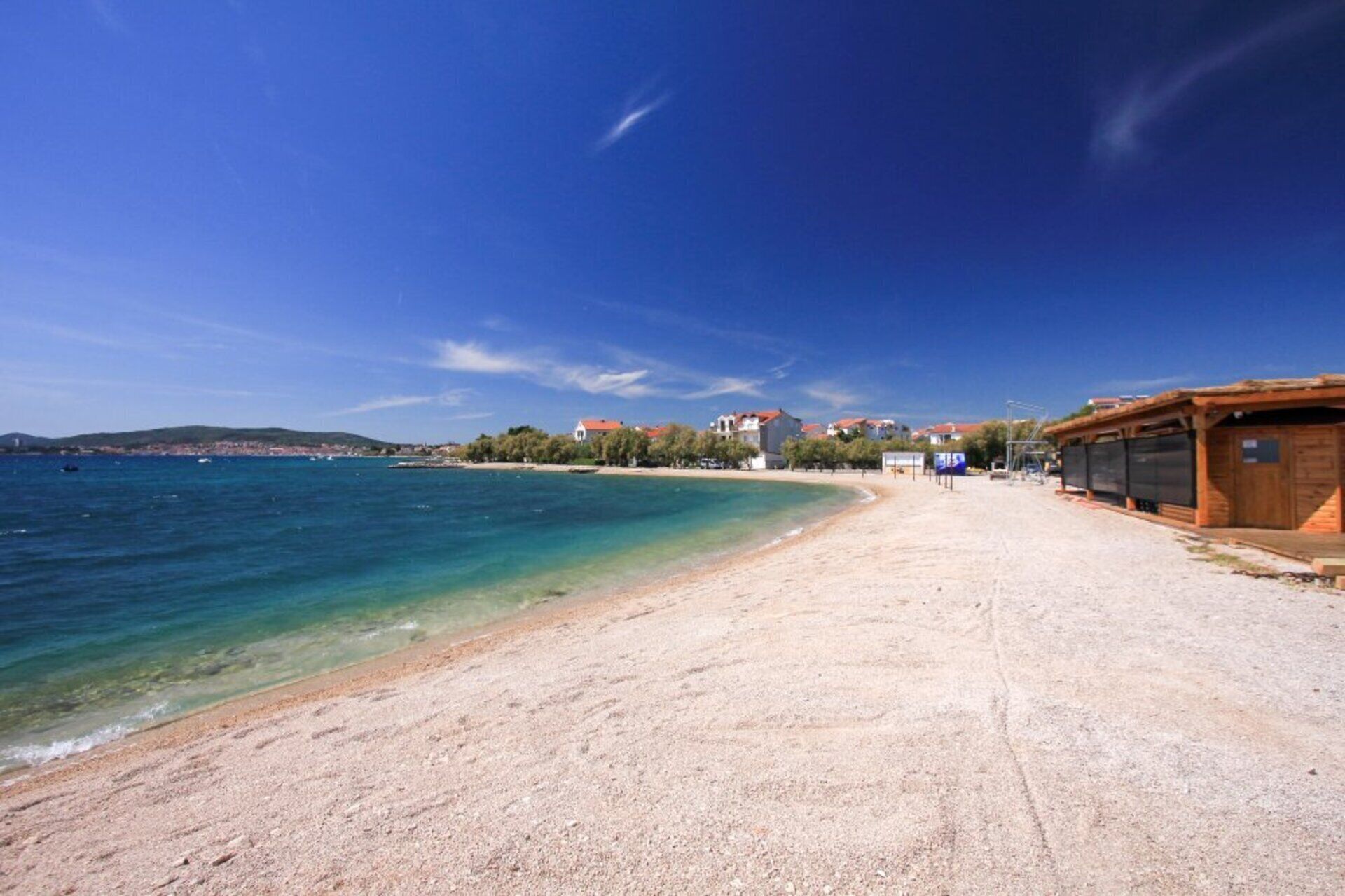 A beach with clear blue water, a sandy shore, and buildings in the background.