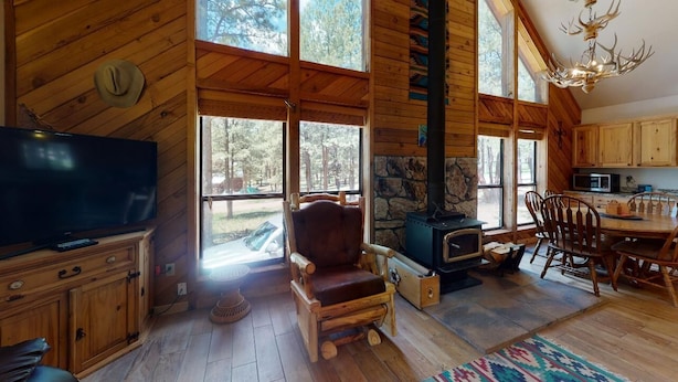 A rustic cabin interior with a stone fireplace, wooden furniture, and large windows offering a view of the forest.