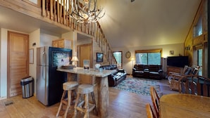 A rustic cabin interior with a wooden staircase, a kitchen area with a refrigerator, a dining table with stools, a sofa, and a television.