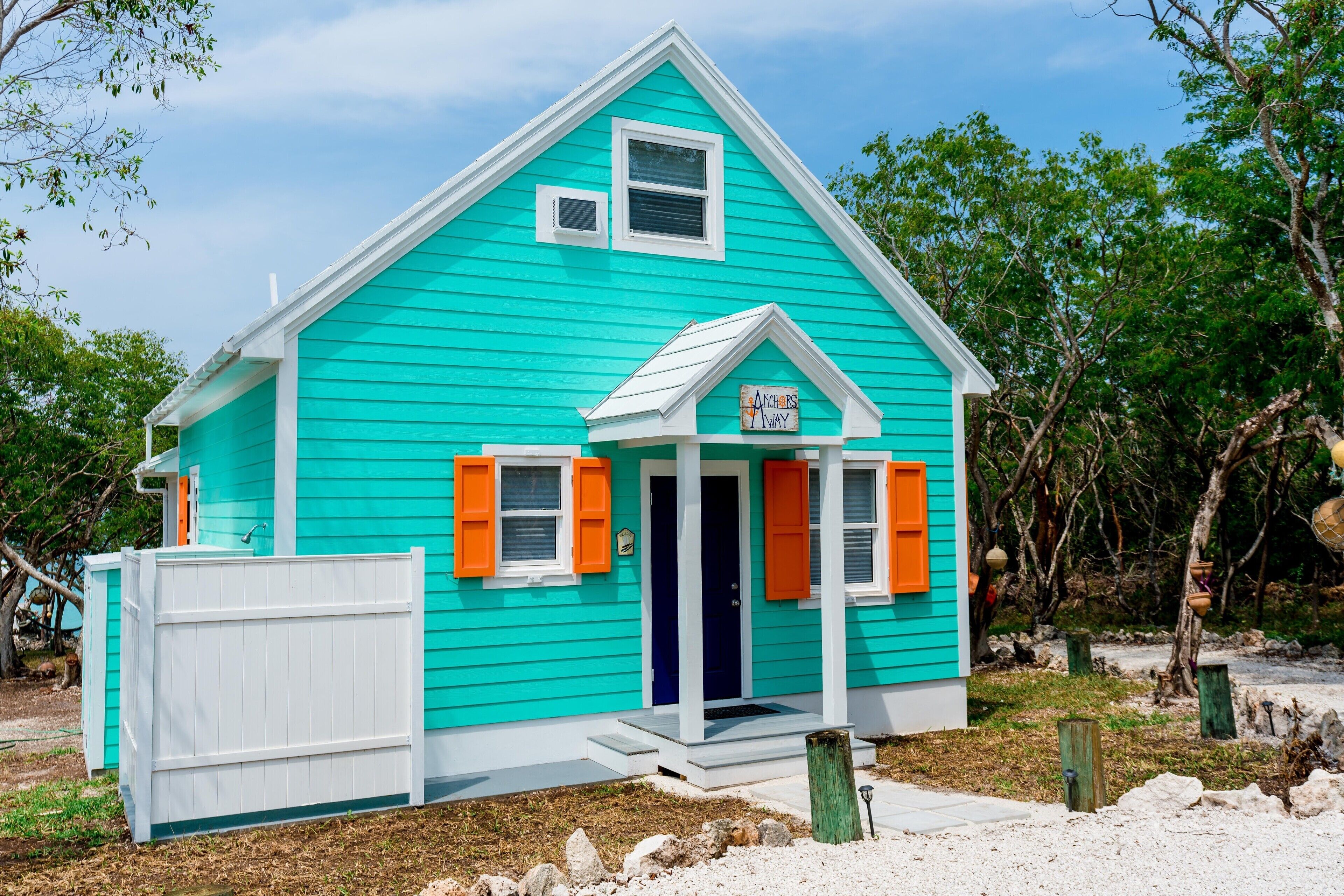 Une maison turquoise avec une porte blanche et des volets orange, entourée d’arbres et d’une clôture blanche.