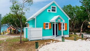 Een turquoise huis met witte details en oranje luiken, omringd door bomen en een zandig gebied.