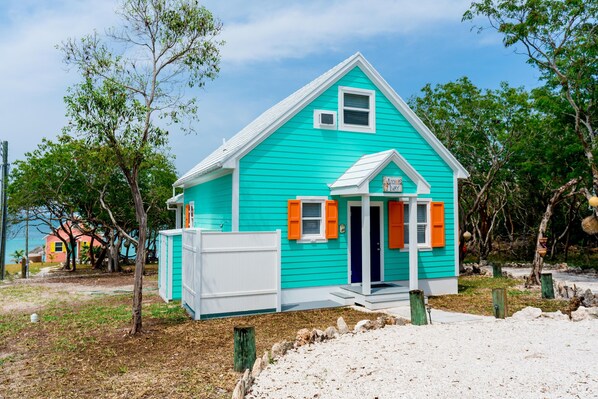 A turquoise house with white trim and orange shutters, surrounded by trees and a sandy area. - Anchor's Away, Russell Island (Russell Island)