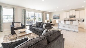 A modern living room with grey sofas, a coffee table, and a dining area with a table and chairs. The kitchen features white cabinets and modern appliances. - Charming Disney Vacation House - The Best Clubhouse (Kissimmee)