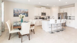 A modern kitchen with white cabinetry, a dining area with a table and chairs, and a cityscape painting on the wall. - Charming Disney Vacation House - The Best Clubhouse (Kissimmee)