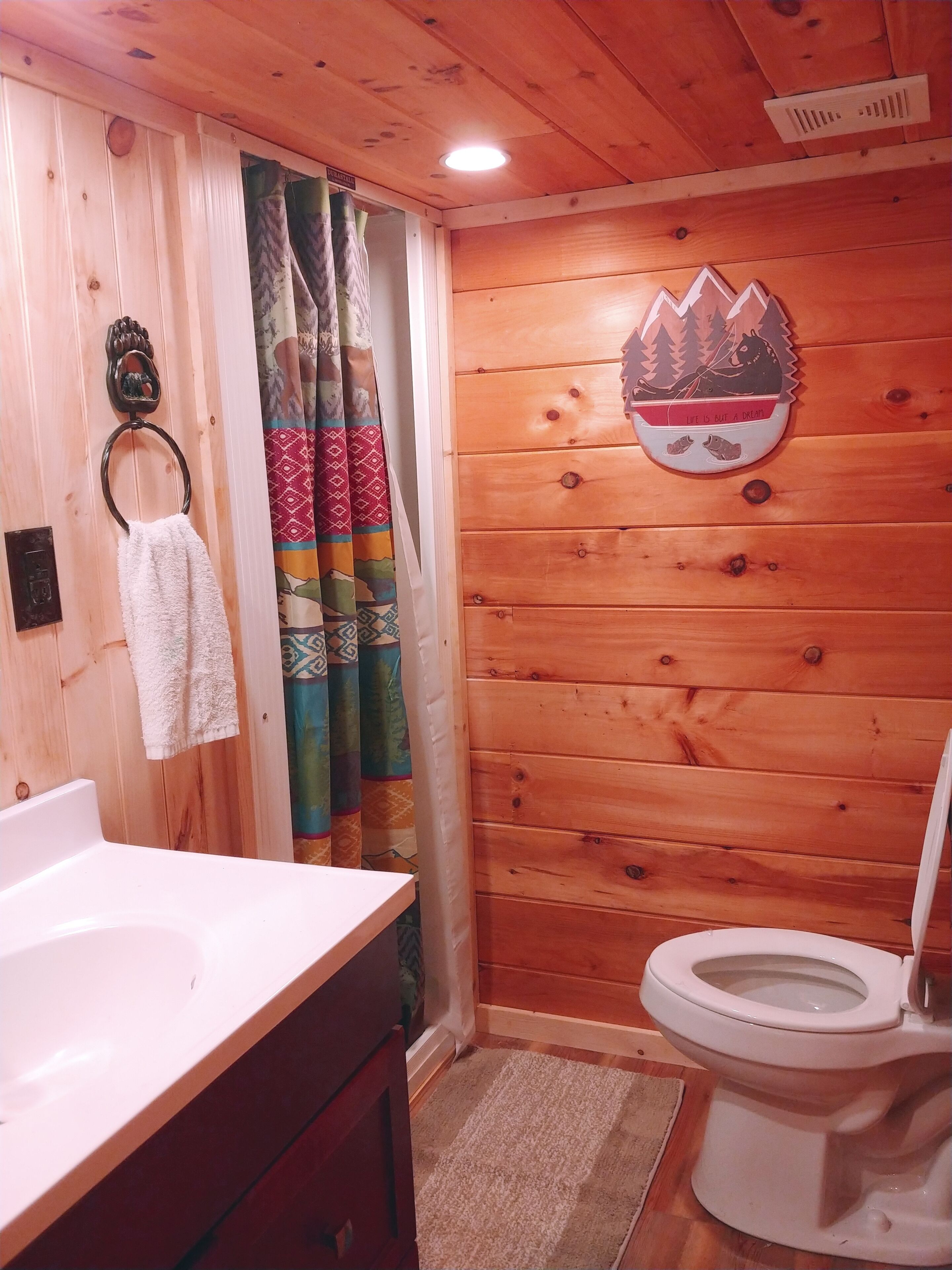 Une salle de bain aux murs lambrissés, avec un lavabo blanc, un porte-serviettes, des toilettes et un rideau coloré.
