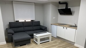 Ett modernt vardagsrum med en grå hörnsoffa, ett vitt soffbord, ett kompakt kök med diskmaskin och en bänkskiva i trä.