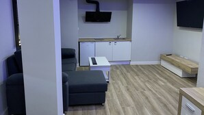 Ett kompakt bostadsutrymme med ett litet kök, en soffa, en TV som är monterad på väggen och ett trägolv.