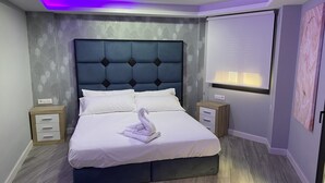 A modern bedroom with a tufted headboard, a swan-shaped towel, a bedside table with drawers, a nightstand, and a wall-mounted TV. - Luxury Gold (Linares)