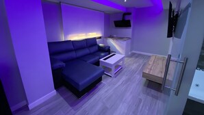 A modern living room with a blue sofa, a small white table, and a kitchen area with a microwave and sink. - Luxury Gold (Linares)