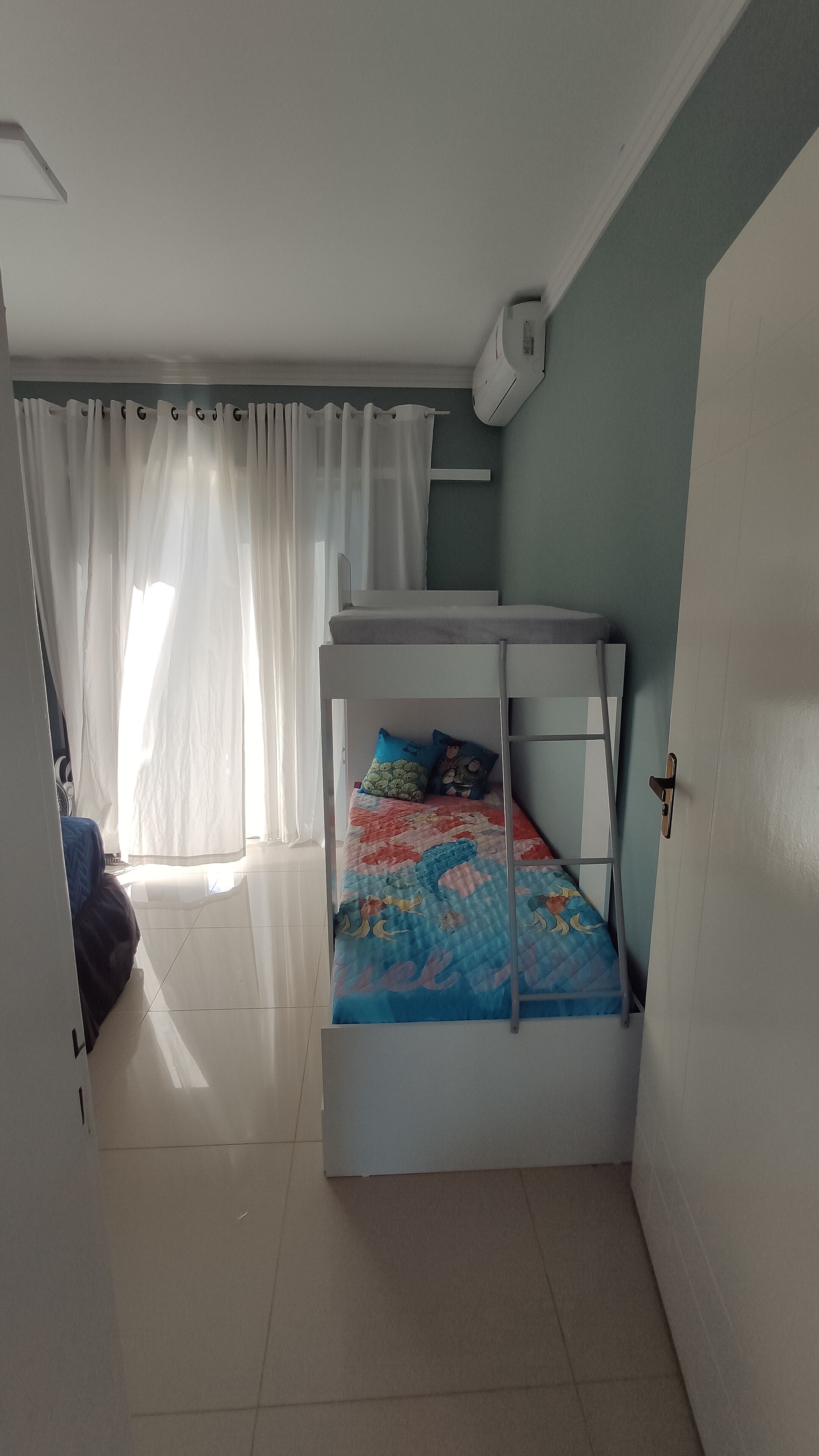 Une petite pièce bien éclairée, avec un lit superposé, une fenêtre ornée de rideaux légers et une porte dotée d’une poignée.