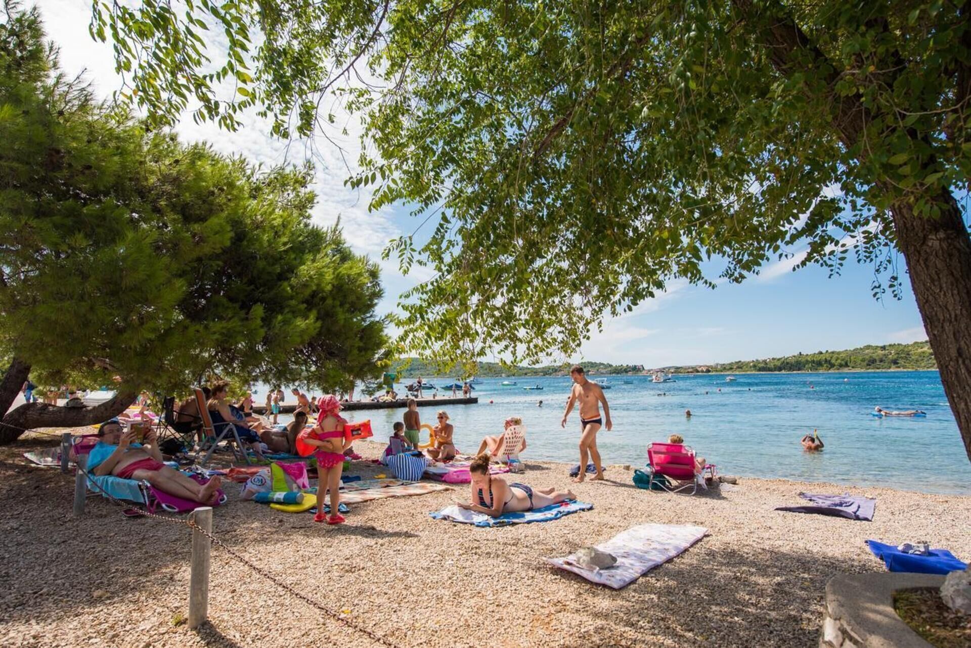 Een strand met mensen die zonnen en zwemmen, omringd door bomen en een heldere lucht.