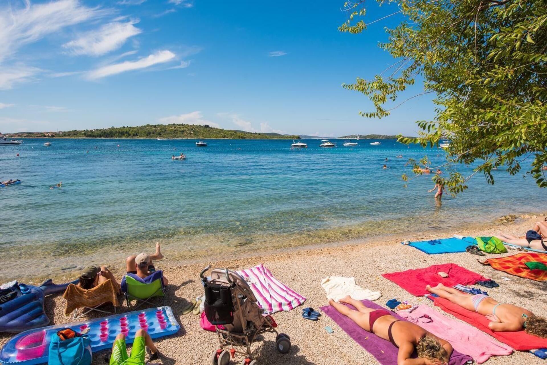 A beach with clear blue water, people sunbathing, and boats in the distance.