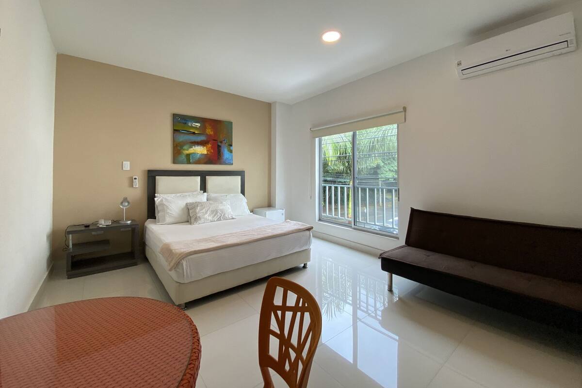 Une chambre d’hôtel équipée d’un lit, d’un bureau, d’une chaise, d’un canapé, d’une fenêtre donnant sur un espace verdoyant et d’un appareil de climatisation.