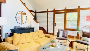 A cozy living room with a yellow sofa, a coffee table, and a fireplace. There is a staircase leading to an upper level, and a door with a window. - The Rocking A Frame - modern meets cozy. (Danbury)