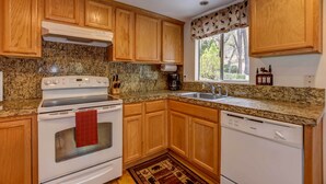 Una cocina con armarios de madera, encimeras de granito, estufa, lavavajillas y ventana con vistas a árboles.