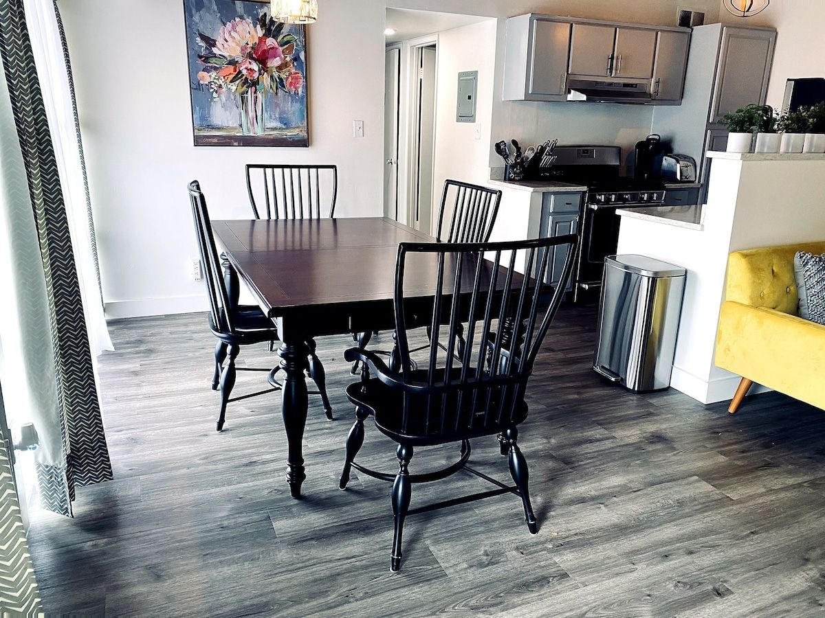 A dining area with a wooden table and chairs, a kitchen in the background, and a yellow sofa.