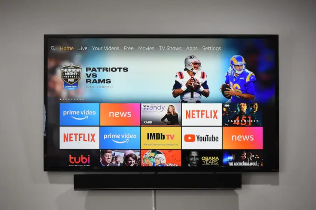 Una pantalla de televisión mostrando un evento deportivo entre los Patriots y los Rams, con varios logotipos de servicios de transmisión visibles.