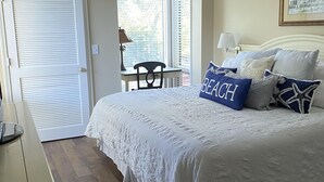 A bedroom with a large bed, a desk, and a chair. There is a window with blinds, a door, and a painting on the wall. - Beautiful Sunsets--Beachfront, Club Amenity Cards included with price! (Seabrook Island)