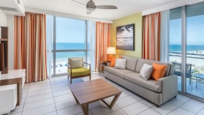 Un salon moderne avec un canapé, un fauteuil et une table basse, de grandes fenêtres avec des rideaux, un ventilateur de plafond et une vue sur la plage.
