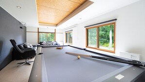 Une table de billard dans une pièce dotée de grandes fenêtres, d’un canapé et d’un plafond avec des poutres en bois.