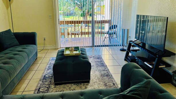 Ein Wohnzimmer mit Sofa, Hocker, Fernseher und Blick durch eine Schiebetür auf eine Terrassenanlage im Freien.