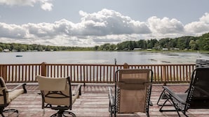 Une terrasse avec des sièges extérieurs donnant sur un lac, un bateau et une rive bordée d’arbres.