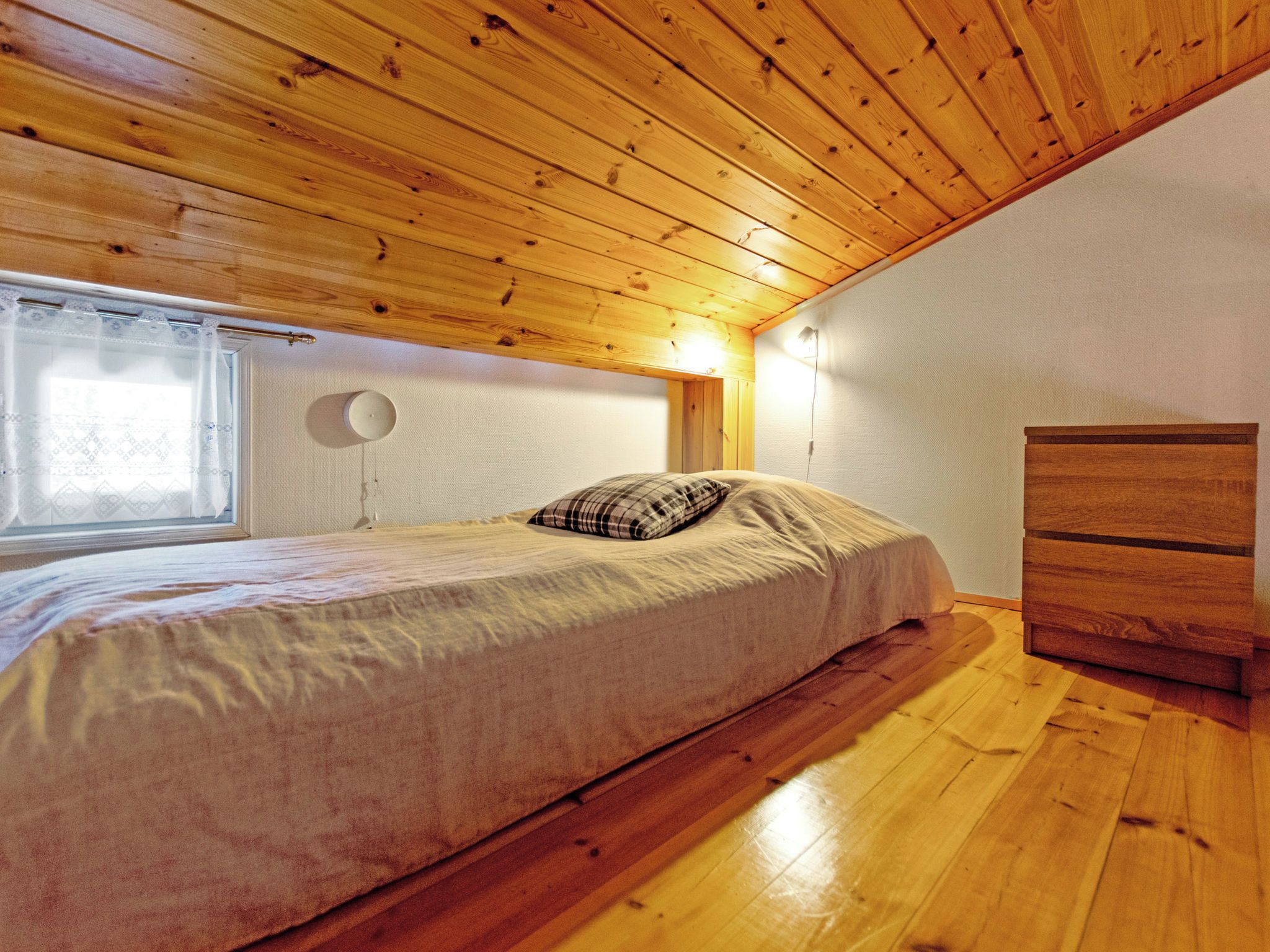 A bedroom with a wooden ceiling, a bed with a plaid pillow, a bedside table, and a dresser.