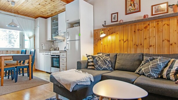 A cozy living room with a sofa, ottoman, and a small table. There is a kitchen area with a refrigerator, oven, and cabinets.